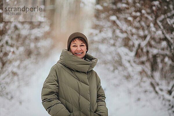 Fröhlich lächelnde junge Frau mit Strickmütze und dunkelgrüner gefütterter Jacke  die im Freien in einer verschneiten Winterwaldlandschaft steht und mit positivem Gesichtsausdruck in die Kamera schaut und das schöne Wetter genießt
