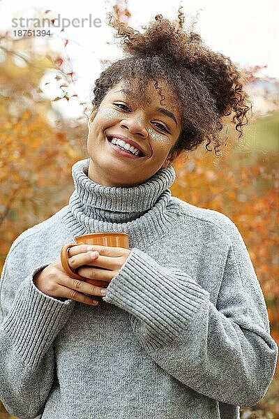 Junge glückliche lächelnde gemischtrassige Frau mit Kaffeetasse in herbstlicher Natur  zufriedene Afroamerikanerin mit lockigem Haar im Strickpullover  die in die Kamera schaut  während sie den Morgen in einem schönen Herbstgarten genießt