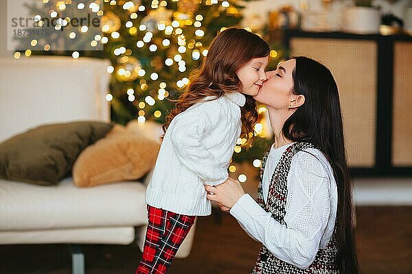 Schöne liebevolle Mutter küsst sanft süße kleine Tochter gegen festlich geschmückten Weihnachtsbaum mit leuchtenden Lichtern auf dem Hintergrund  glückliche Familie verbringen Silvester zu Hause zusammen
