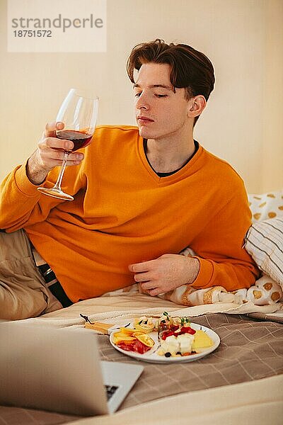 Entspannter junger Mann mit einem Glas Wein in der Hand  der auf einem bequemen Bett mit einem Teller Essen liegt und beim Essen Videos auf dem Laptop anschaut  während er sich zu Hause entspannt