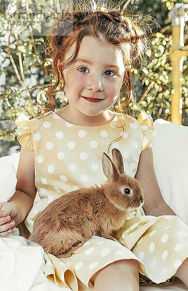 Kleines hübsches Mädchen im Sommerkleid sitzt draußen auf dem Bett mit Kaninchen  niedliche Kind Blick auf Hasen mit leichten Lächeln genießen Zeit mit ihren Lieblings Tiere