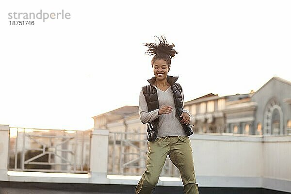 Junge glückliche sorglose afrikanische amerikanische Frau tanzt und hat Spaß auf dem Dach im morgendlichen Sonnenlicht mit Stadtlandschaft im Hintergrund  glückliche gemischtrassige Frau in Freizeitkleidung  die sich auf dem Dach zur Musik bewegt
