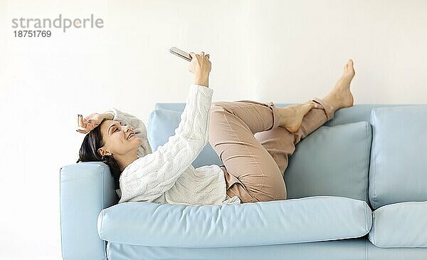 Junge brünette Frau liegt auf einem gemütlichen Sofa in entspannter Position  die Beine hochgeworfen  hält ihr Handy  schaut aufmerksam auf den Bildschirm  chattet online in ruhiger häuslicher Umgebung  liest wichtige Nachrichten