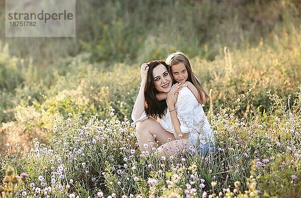 Lächelnde Mutter und Tochter  die sich auf einer blühenden Wiese aneinander kuscheln  während sie ein Wochenende auf dem Lande verbringen und in die Kamera schauen