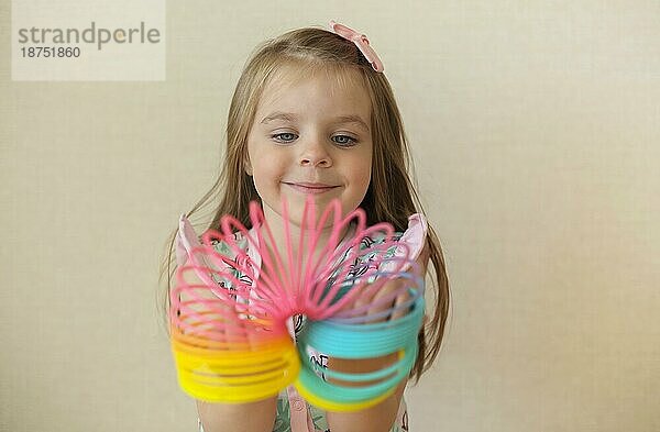 Cute adorable kleines Mädchen spielt mit Kunststoff Regenbogen magische Feder  hält bouncy dehnbar Spielzeug und lächelt in die Kamera. Glückliches Kind vorbeige Hintergrund genießen Freizeit zu Hause