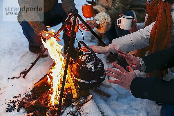 Eine Gruppe von Freunden versammelt sich um ein Lagerfeuer im Hinterhof  trinkt Tee und wärmt sich die Hände. Zwei glückliche Paare  die sich entspannen und die Wintersaison genießen  während sie am Feuer sitzen. Outdoor Winter unterhaltsam