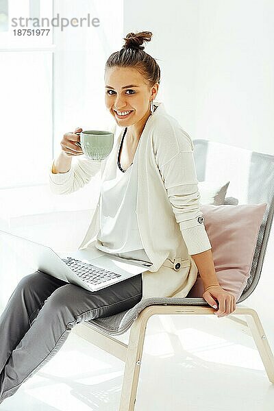 Barfüßige junge Frau sitzt auf einem Stuhl und entspannt sich mit einer Tasse Kaffee  nachdem sie einen Laptop in einem hellen Raum zu Hause benutzt hat