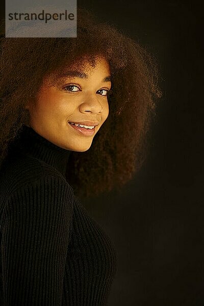 Porträt einer attraktiven  lächelnden  lockigen afroamerikanischen Frau in schwarzer Kleidung  die in die Kamera schaut und vor einem dunklen Studiohintergrund posiert. Junges natürlich schönes afrikanisches weibliches Modell ohne Make up