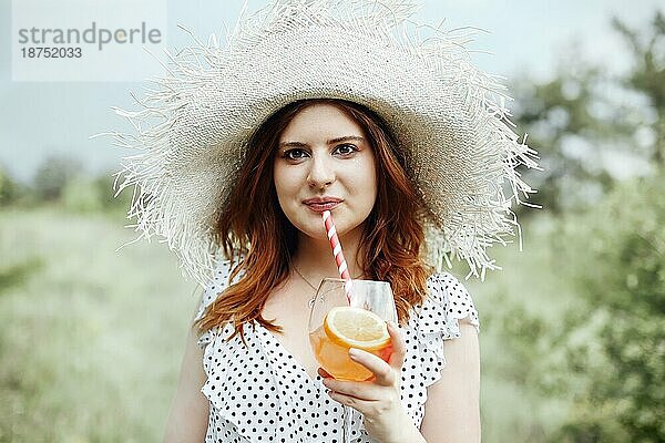 Junge Frau mit Sommerhut lächelt und schaut in die Kamera  während sie einen Alkoholcocktail mit Orange auf einem unscharfen Hintergrund in der Natur trinkt