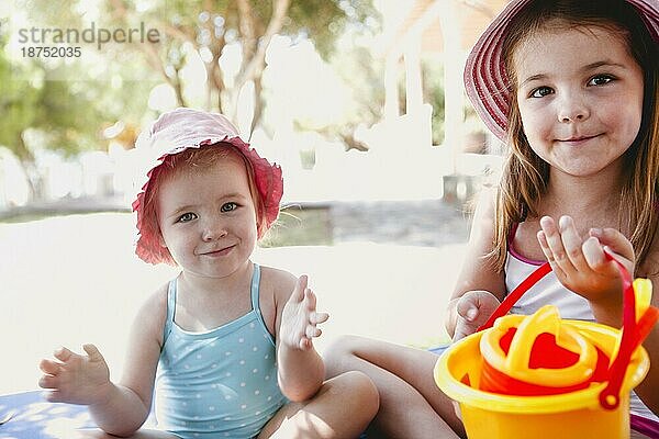Bezaubernde Schwestern in Sommerkleidung sitzen im Schatten am Ufer und spielen mit Plastikspielzeug