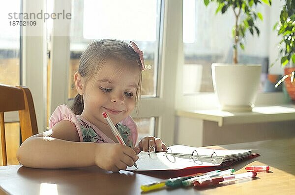 Adorable lächelnd kleines Mädchen sitzt am Tisch und Zeichnung mit multicolor Gel Stifte in Notebook  glücklich Kind genießen Kunst Aktivität während der Freizeit zu Hause. Preschooler kreatives Lernen und Entwicklung