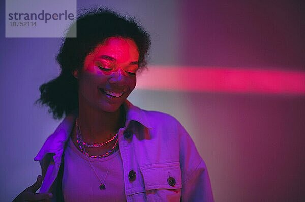 Junge glückliche fröhliche afrikanische amerikanische Frau  die unter neonleuchtendem Licht in einem Studio steht und sanft lächelt  gekleidet in Denim Freizeitkleidung