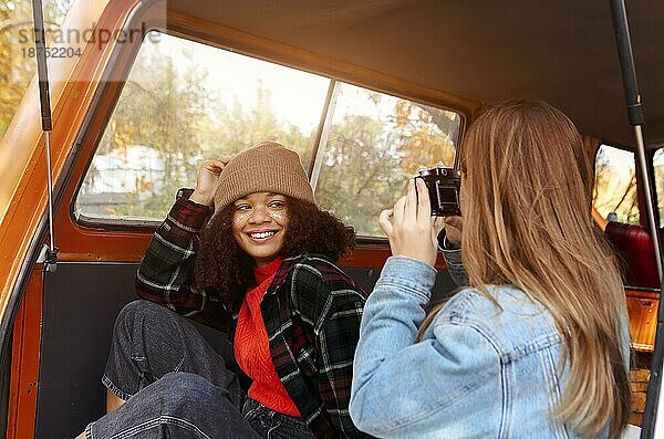 Junge lächelnde afroamerikanische Frau  die für ein Foto posiert  während sie mit ihrer Freundin auf dem Kofferraum eines Autos sitzt  zwei verschiedene multirassische Freundinnen in einem Minivan  die ein Fotoshooting im Herbstwald machen
