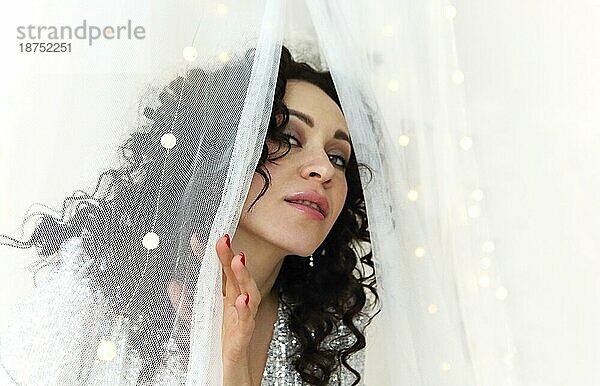 Close up von jungen sinnlichen attraktive Frau mit lockigem Haar tragen festliche Make up schaut aus hinter weißen Vorhang mit xmas führte Lichter dekoriert. Weibliche Schönheit Konzept