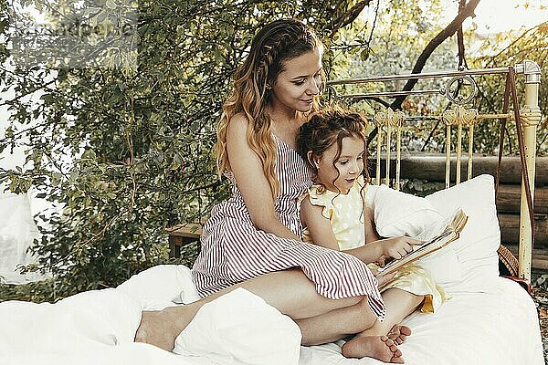Schöne Mutter und Kind verbringen Zeit zusammen auf sonnigen Terrasse in der Nähe von Haus  Mama liest Märchen laut zu reizenden Mädchen  während kleines Mädchen sitzt auf ihrem Schoß Sommerferien wof die Familie