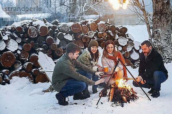 Eine Gruppe von Freunden versammelt sich um ein Lagerfeuer im Hinterhof  trinkt Tee und wärmt sich die Hände. Zwei glückliche Paare  die sich entspannen und die Wintersaison genießen  während sie am Feuer sitzen. Outdoor Winter unterhaltsam