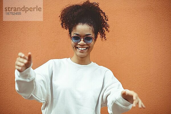 Junge Frau afrikanischer Abstammung mit stilvoller Sonnenbrille  mit lockigem Haar  das zu einem hohen Pferdeschwanz gebunden ist  schaut weg  während sie breit lächelt und gerade  perfekte Zähne zeigt  und posiert vor einem orangefarbenen Wandhintergrund