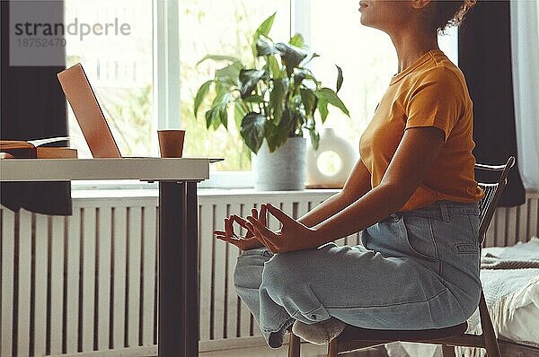 Stressabbau während der freiberuflichen Arbeit. Cropped Foto von afrikanischen amerikanischen Frau Freiberufler sitzen in Lotuspose und meditieren mit den Händen in Mudra Geste auf Stuhl vor dem offenen Laptop auf dem Tisch