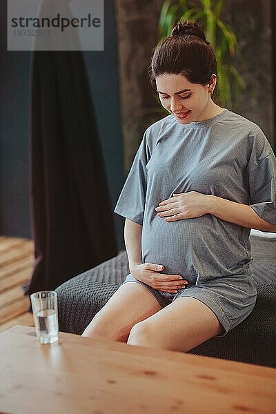 Gesunde Gewohnheiten während der Schwangerschaft. Junge glückliche schwangere Frau in Homewear berührt sanft den Bauch  während sie am Tisch mit einem Glas reinen Wassers sitzt und Wasser trinkt  um ihren Körper und ihr Baby gesund zu halten