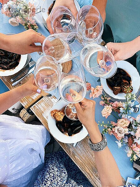 Gruppe von Freunden  die mit Gläsern Weißwein über einem Tisch mit mediterranen Muscheln jubeln  während sie ein Abendessen im Freien genießen  Menschen stoßen beim Sommerpicknick mit Weingläsern an  Blick von oben
