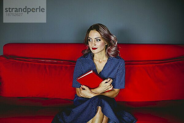 Elegante Frau mit roten Lippen im Retrokleid sitzt auf dem Sofa und genießt ein interessantes Buch