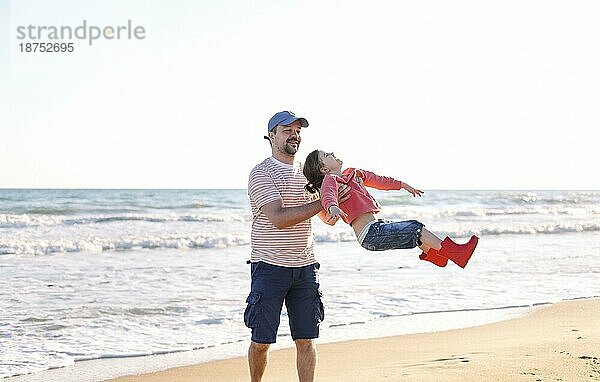 Liebevoller Vater spielt mit seiner entzückenden kleinen Tochter am Strand  schaukelt sie herum  Vater und Kind verbringen Zeit im Freien zusammen  sonniges Meer im Hintergrund. Parenting Konzept