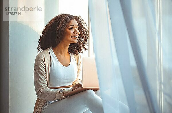 Fröhliche afroamerikanische junge Frau mit lockigem Haar sitzt auf der Fensterbank mit Laptop auf dem Schoß  tippt auf der Tastatur  schaut auf den Bildschirm und lächelt  glückliche afrikanische Frau  die mit Freunden online chattet