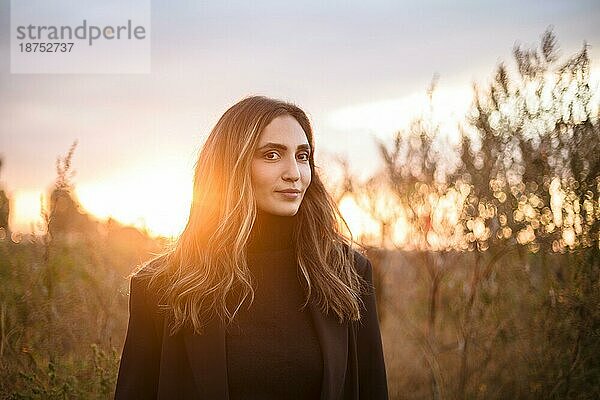 Junge glückliche kaukasische brünette Frau  die bei Sonnenuntergang über eine Wiese läuft und lächelt  verträumte schöne Frau im Abendsonnenlicht  die auf einem herbstlichen Feld auf dem Lande steht