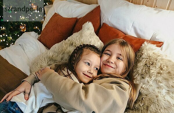 Gemütliche inländischen Porträt von zwei glücklichen lächelnden kleinen Mädchen Schwestern umarmt  während die Freizeit zusammen zu Hause zu verbringen  auf dem Bett in der Nähe geschmückten Weihnachtsbaum liegen. Familie und Kinder Konzept