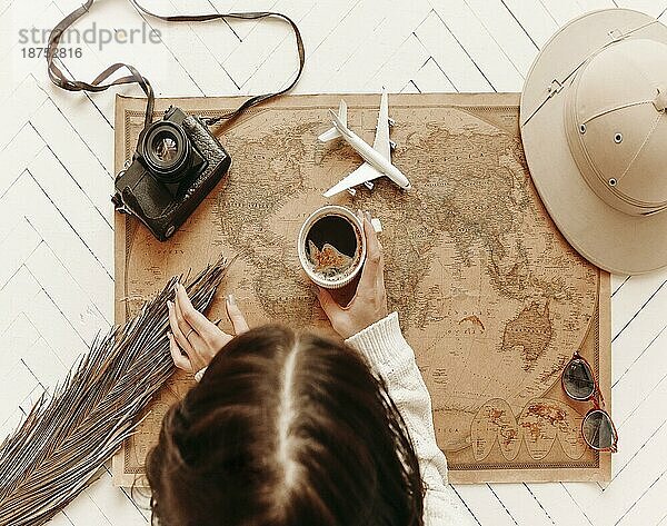 Ansicht von oben von weiblichen hält Tasse schwarzen Kaffee über Vintage Look Weltkarte mit Reisenden Zubehör  Retrokamera Spielzeugflugzeug auf der Oberseite des Parkettbodens. Tourismus und Urlaub Konzept