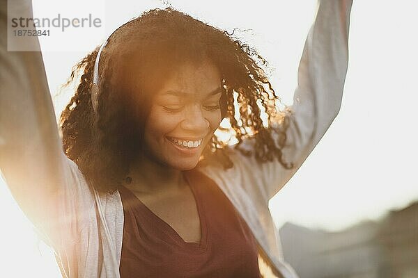 Ganzkörperaufnahme einer jungen  sorglosen afroamerikanischen Frau  die auf einem Dach im morgendlichen Sonnenlicht springt  mit einer Stadtlandschaft im Hintergrund  eine glückliche  gemischtrassige Frau in Freizeitkleidung  die das Leben und den neuen Tag genießt