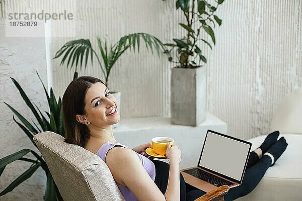 Glückliche schwangere junge erwachsene Frau sitzt auf einem Sofa  arbeitet an einem Laptop und hält eine Tasse Kaffee. Schwangerschaft zu Hause Konzept