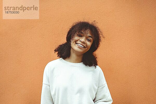 Pures Glück. Nettes glückliches afroamerikanisches Mädchen mit lockiger Frisur und weißem Pullover posiert gegen eine orangefarbene Wand. Lächelnde schwarze junge Frau  die mit ihrem Leben zufrieden ist