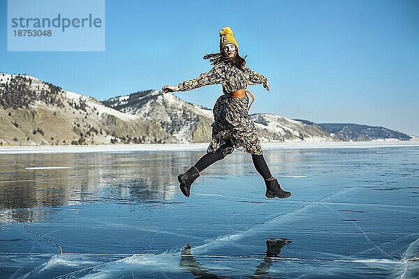 Modisches Bild einer atemberaubenden Frau  die auf dem Eis des Baikalsees posiert und einen gelben Hut trägt. Boho Kleid fliegt im Wind. Schöne Eislandschaften des Baikalsees