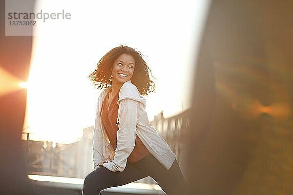 Junge sportliche gemischtrassige Frau macht Aufwärm Stretching Übungen und lächelt  während sie am Morgen auf dem Dach steht  mit einer Stadtlandschaft im Hintergrund  eine fröhliche afroamerikanische Frau  die bei Sonnenaufgang trainiert