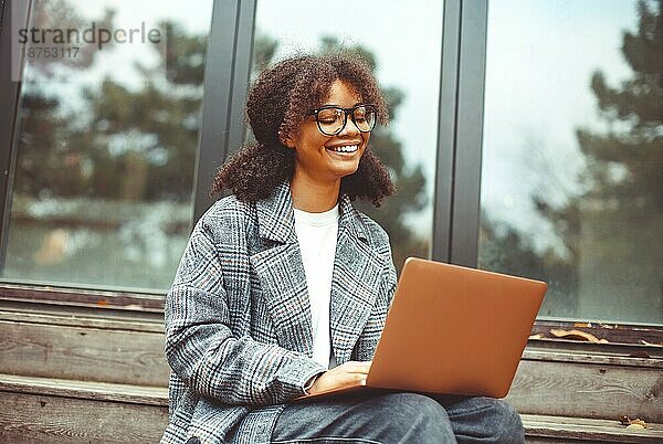 Outdoor Herbst Porträt der jungen fröhlichen afrikanischen amerikanischen Frau mit Laptop auf den Knien sitzen auf hölzernen Haus Treppe  positive schwarze Mädchen in Brillen arbeiten remote in der Natur auf Herbsttag