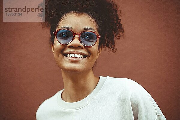 Junge Frau afrikanischer Abstammung mit stilvoller Sonnenbrille  mit lockigem Haar  das zu einem hohen Pferdeschwanz gebunden ist  schaut weg  während sie breit lächelt und gerade  perfekte Zähne zeigt  vor einem braunen Wandhintergrund posierend