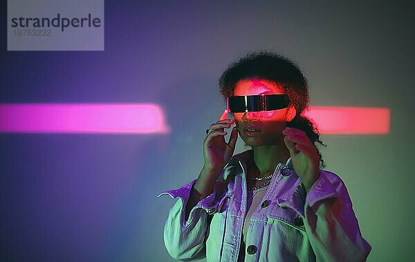Junge selbstbewusste afroamerikanische Frau mit VRheadset die die virtuelle Welt erkundet  während sie in Jeans und Freizeitkleidung unter Neonlicht im Studio steht und Virtual Reality Spiele spielt. Moderne Technologien