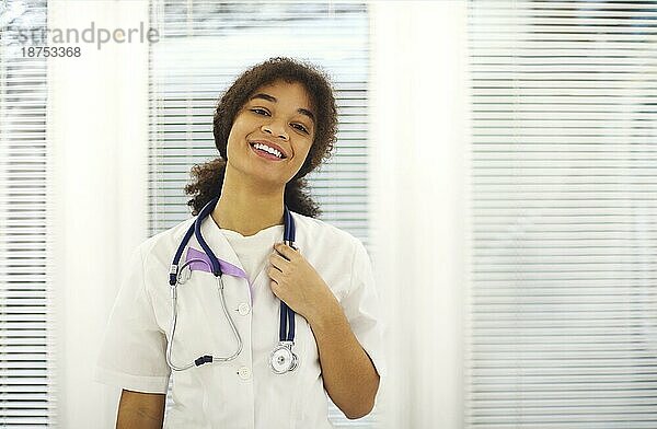 Glückliche afroamerikanische Ärztin in medizinischer Uniform mit Stethoskop um den Hals lächelt in die Kamera während der Arbeitsschicht in einer modernen Klinik