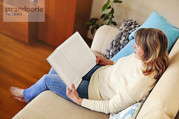 Von oben Seitenansicht der ruhigen reifen Frau in lässiger Kleidung sitzt auf bequemen Sofa und Lesung Buch beim Ausruhen zu Hause