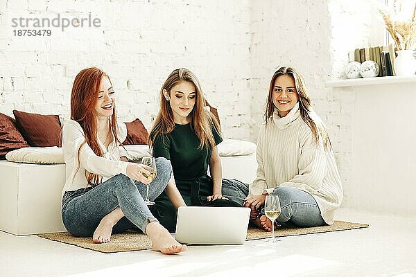 Fröhliche junge Frauen mit Weingläsern  die lächelnd auf ihrem Netbook surfen  während sie sich zu Hause auf dem Boden neben der Couch ausruhen