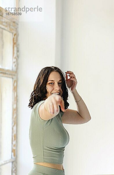 Foto in Bewegung von fröhlicher junger Frau  die springt oder Sport in der Halle treibt  mit breitem Lächeln in die Kamera schaut  während sie sich bewegt  und vor einem unscharfen Wohnungshintergrund posiert