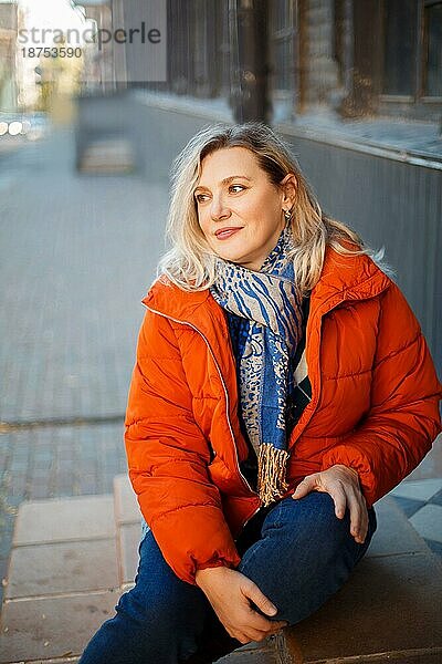 Glücklich lächelnde Frau mittleren Alters in orangefarbener Daunenjacke  die auf einer Betontreppe im Freien sitzt und mit positivem Gesichtsausdruck in die Kamera blickt  während sie sich an einem kalten Herbsttag in der Stadt ausruht