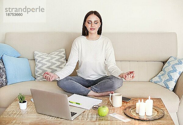 Crop junge Frau in Freizeitkleidung meditieren in Lotuspose beim Sitzen auf dem Sofa vor dem Tisch mit Laptop und brennenden Kerzen in gemütlichen Wohnzimmer zu Hause