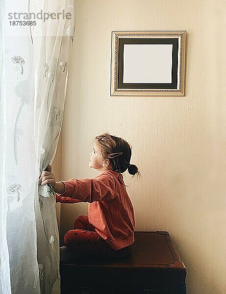 Seitenansicht eines niedlichen kleinen Mädchens  das am Fenster sitzt und glücklich lächelt  während es seine Freizeit zu Hause verbringt  ein kleines Kind  das durch das Fenster schaut und draußen sein möchte oder auf seine Eltern wartet  die gerade von der Arbeit kommen