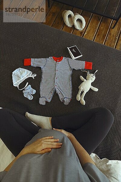 Cropped Schuss der schwangeren Frau Blick auf niedliche Babykleidung während auf dem Bett zu Hause ausruhen  weibliche werdendes Kind genießt Schwangerschaft  die Vorbereitung für die Elternschaft und Geburt. Adorable neugeborenen Kleidung