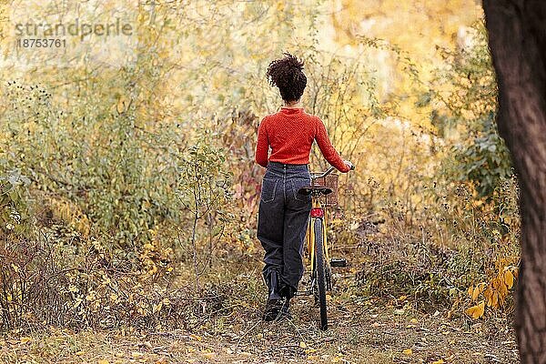 Wochenendaktivität in der Herbstzeit. Junge glückliche lächelnde gemischtrassige Frau mit Fahrrad im Herbst Wald. Rückenansicht der positiven aktiven afrikanischen amerikanischen Frau in Freizeitkleidung Reiten Stadt Fahrrad in der Natur