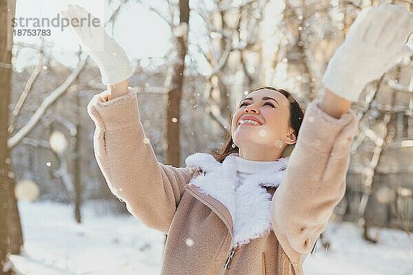 Optimistische junge Frau in modischem Mantel  die lächelnd nach oben schaut und Schneeflocken auffängt  während sie sich am Wochenende auf der Straße amüsiert