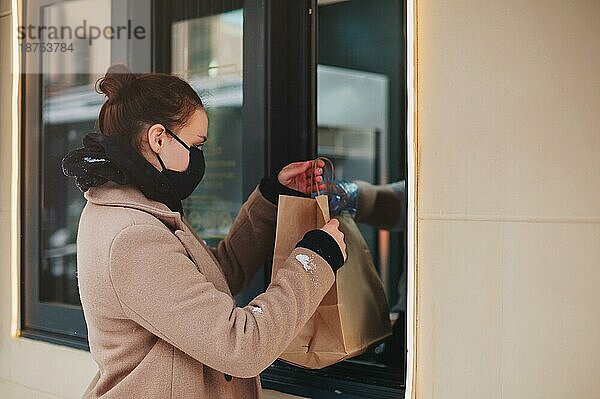 Seitenansicht einer Kundin mit Maske  die eine Papiertüte mit Essen zum Mitnehmen aus dem Fenster eines Diners nimmt  während einer Pandemie