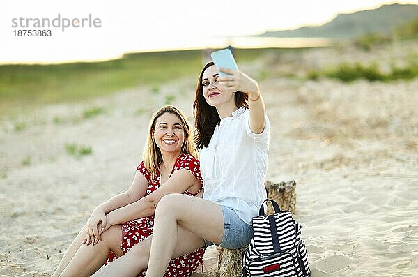 Fröhliche Freundinnen sitzen am Sandstrand und machen ein Selfie mit ihrem Smartphone im Sommer